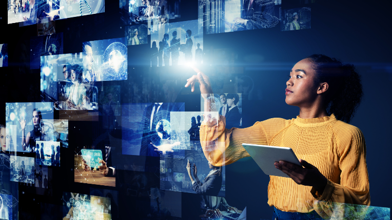Woman holding tablet while standing in front of many digital images; she is pointing at one