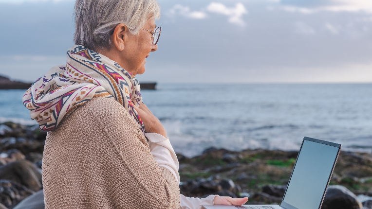 Senior active woman sitting on the pebble beach using laptop in remote work. Mature female enjoying sunset at sea, holding laptop on knees, horizon over water.