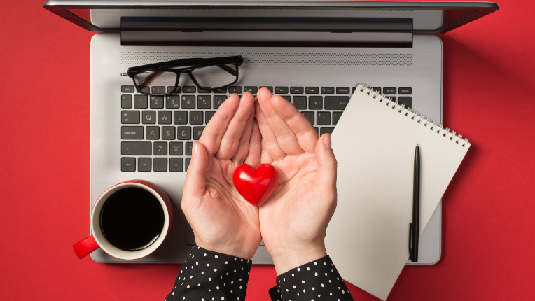Overhead photo of grey laptop notepad pen glasses cup of coffee and hands holding a red heart isolated on the red backdrop.