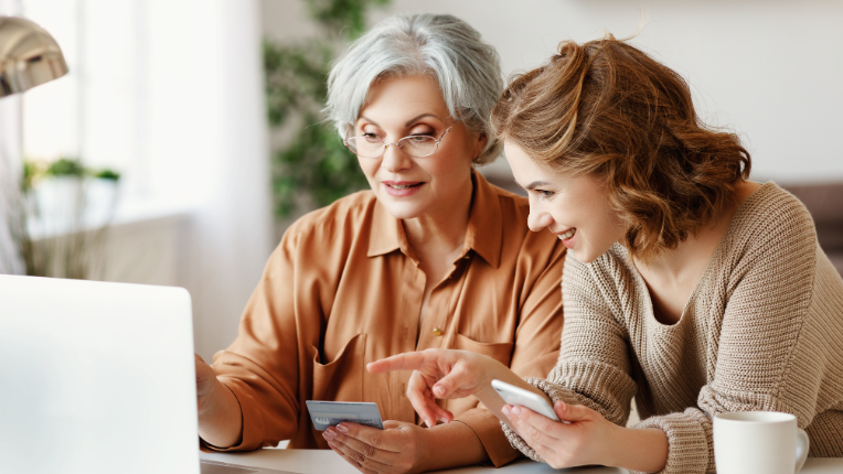 Young woman helping senior woman (holding bank card) use laptop for online banking.
