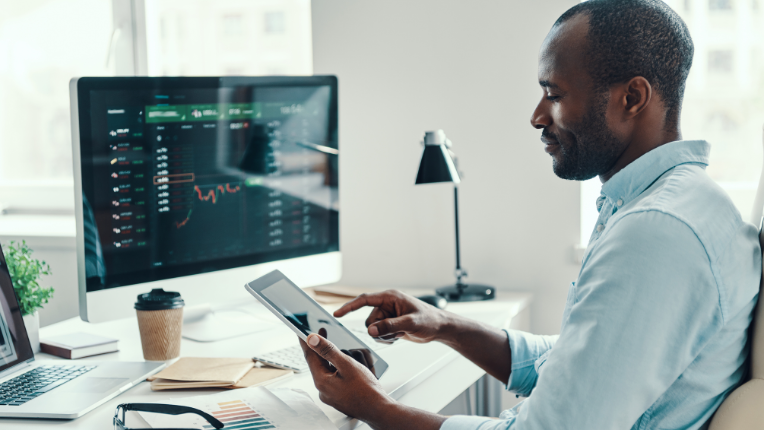 Man sitting at desk looking at his tablet with laptop and computer monitors displaying graphs.